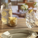 Glass of water with ice cubes and lemon slice on table, with pitcher and lemon wedges in the background.